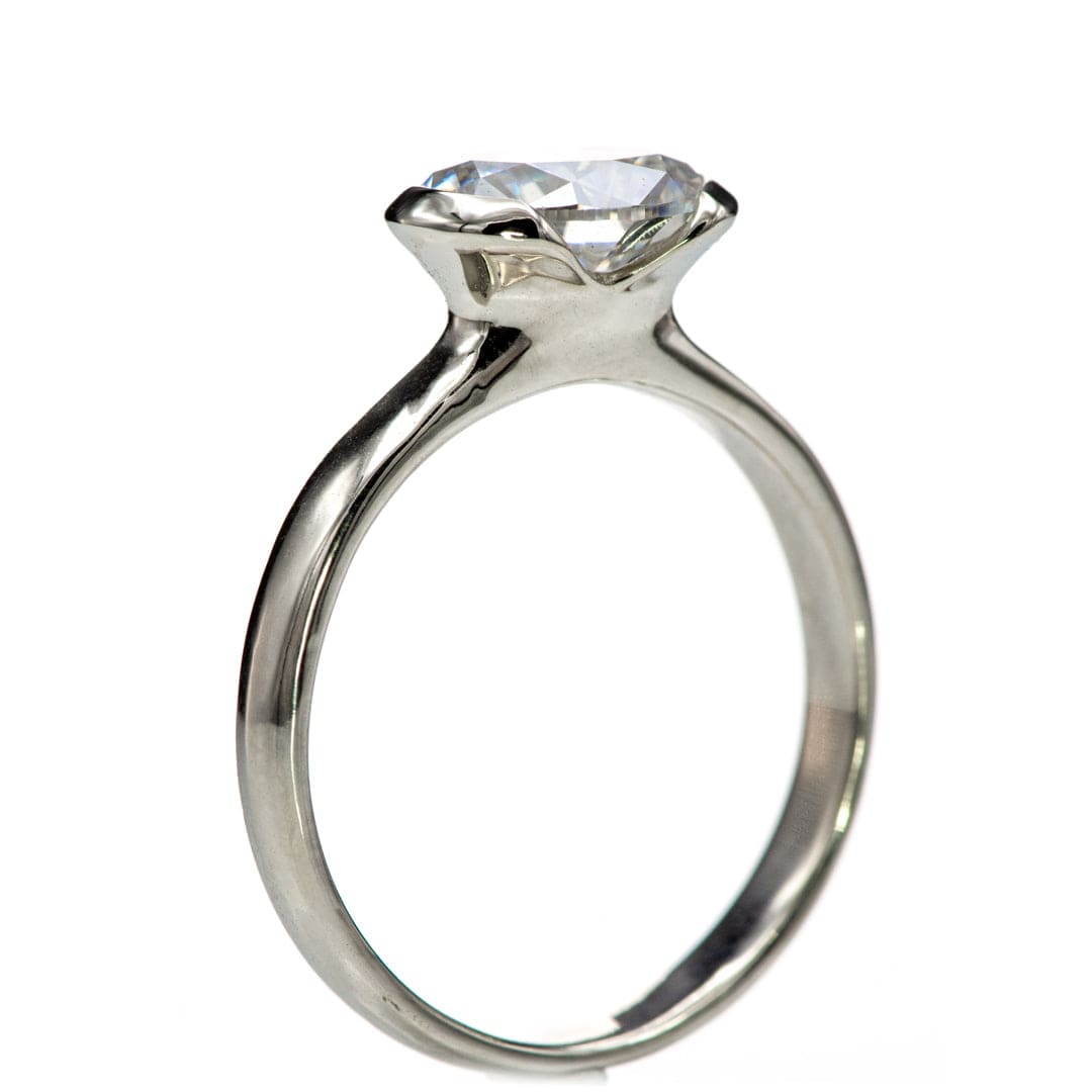 A white gold ring with a single oval cut clear Moissanite gemstone mounted in a semi-bezel setting, viewed from a side angle against a white background. Ring by Nodeform