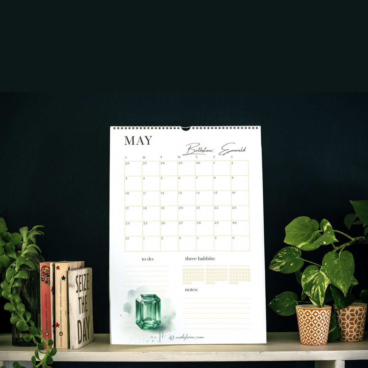2026 Calendar page for May on a shelf with plants and books against a dark background