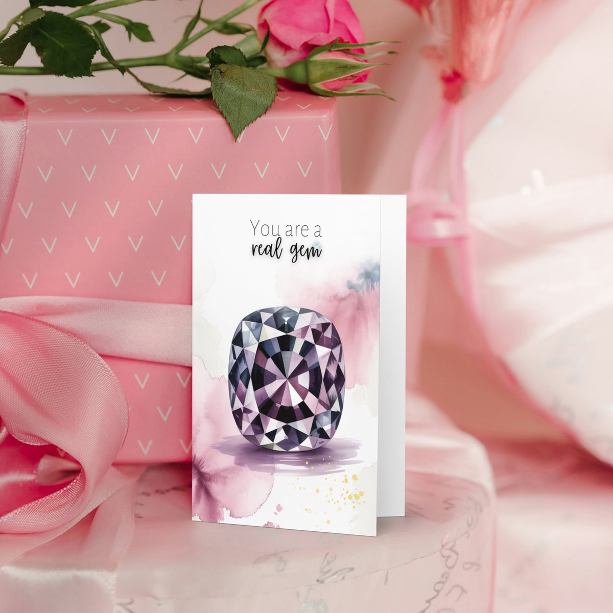 Greeting card with a cushion spinel watercolor illustration and 'You are a real gem' text, placed on pink fabric with roses.