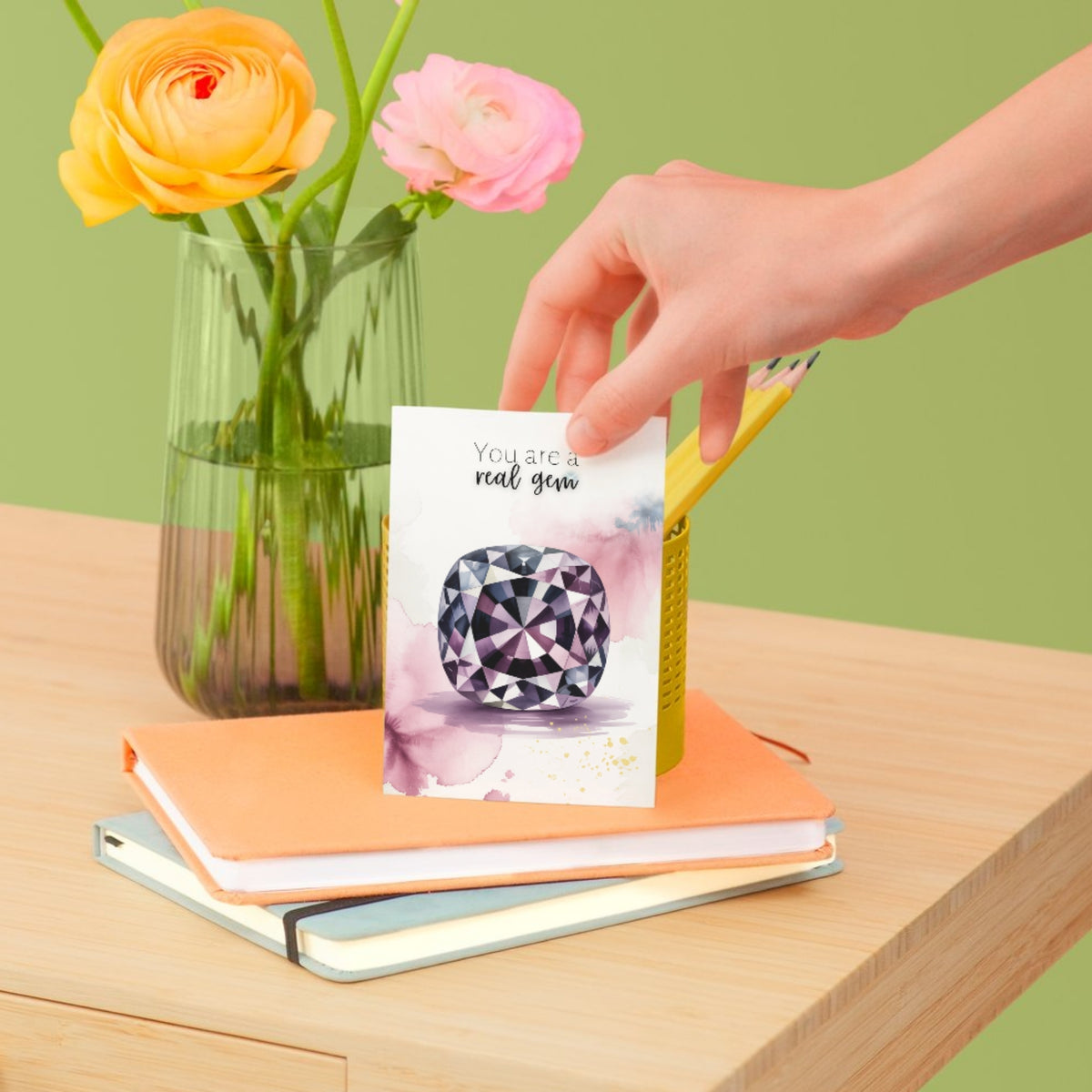 Hand placing a card with a diamond design on books next to a vase of flowers on a wooden surface.