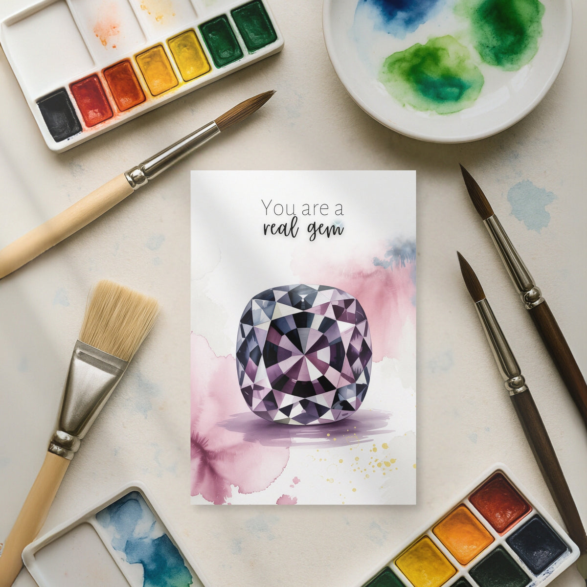 Card with watercolor cushion spinel illustration surrounded by art supplies on a light surface