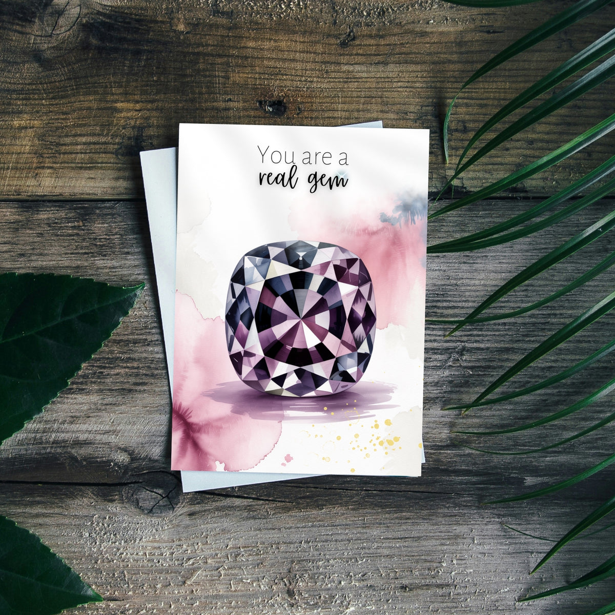 Greeting card with a spinel watercolor illustration and text 'You are a real gem' on a wooden surface with leaves.