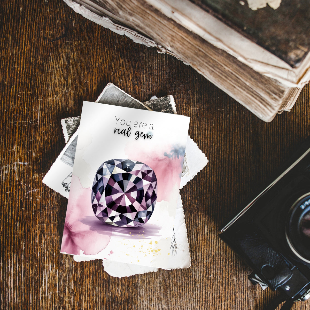 Card with a cushion spinel illustration and 'You are a real gem' text on a wooden surface with a camera and book.