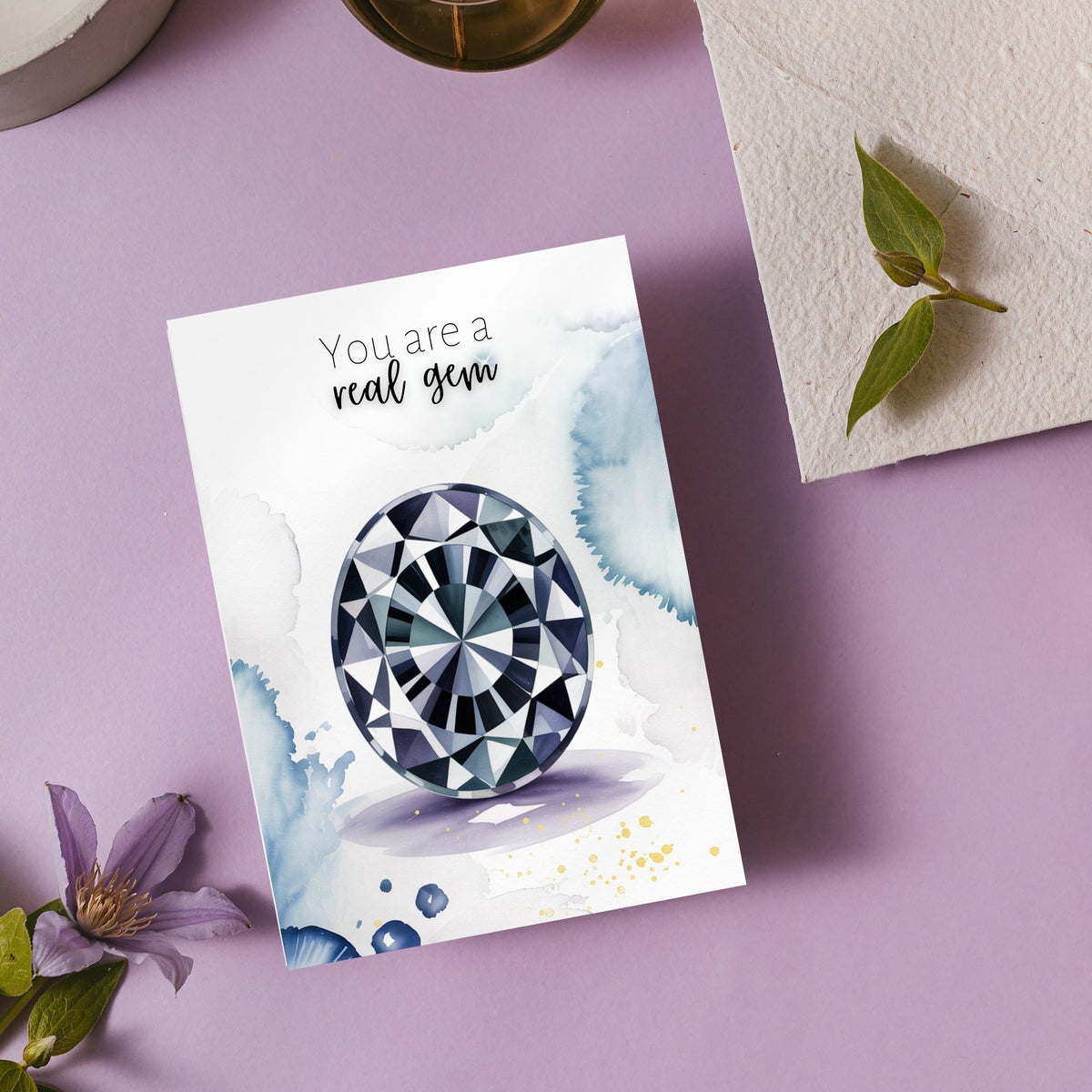 Greeting card with oval Spinel Gemstone illustration and text 'You are a real gem' on a purple surface with flowers and leaves.