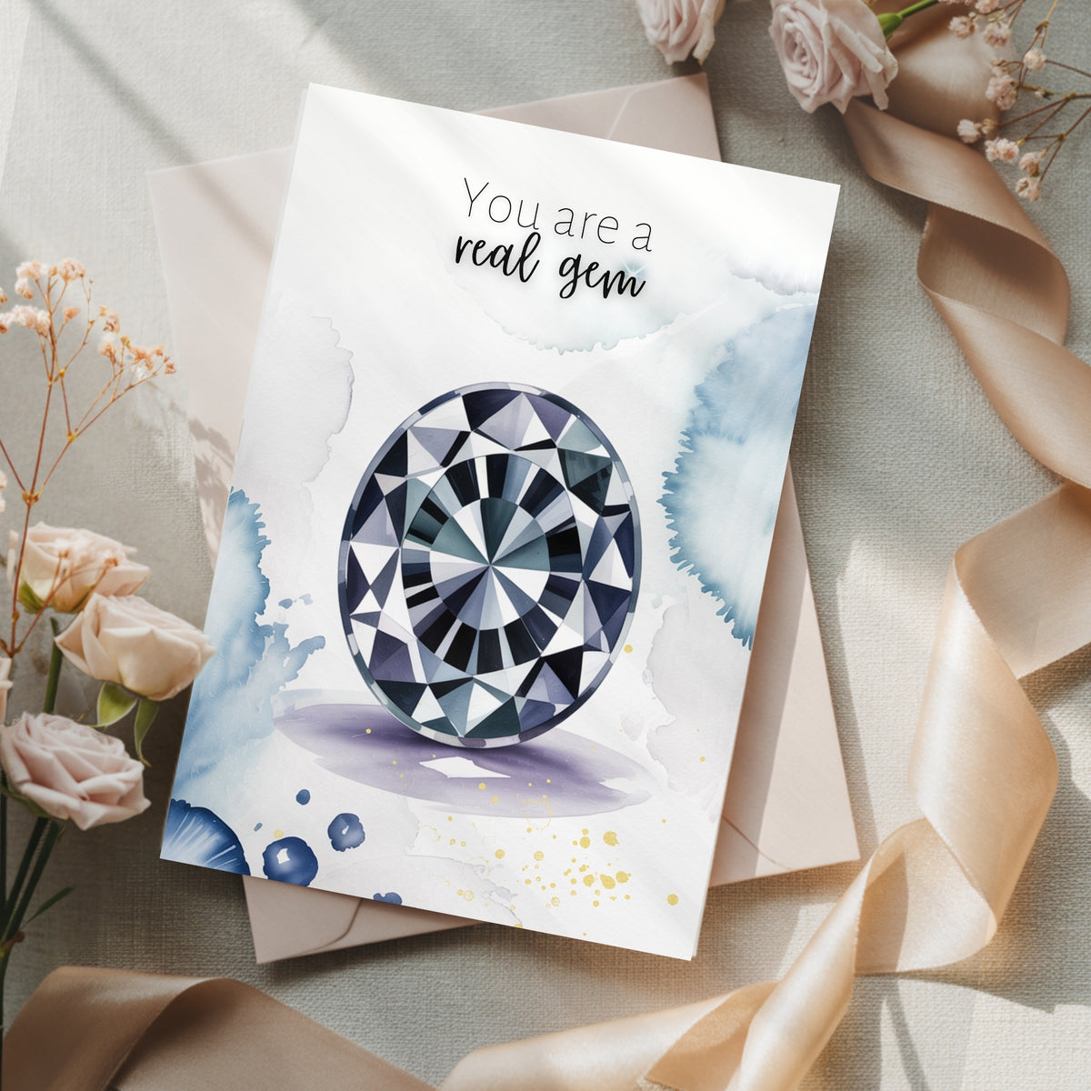 Greeting card with oval Spinel watercolor illustration and 'You are a real gem' text on a decorative surface with flowers and ribbons.