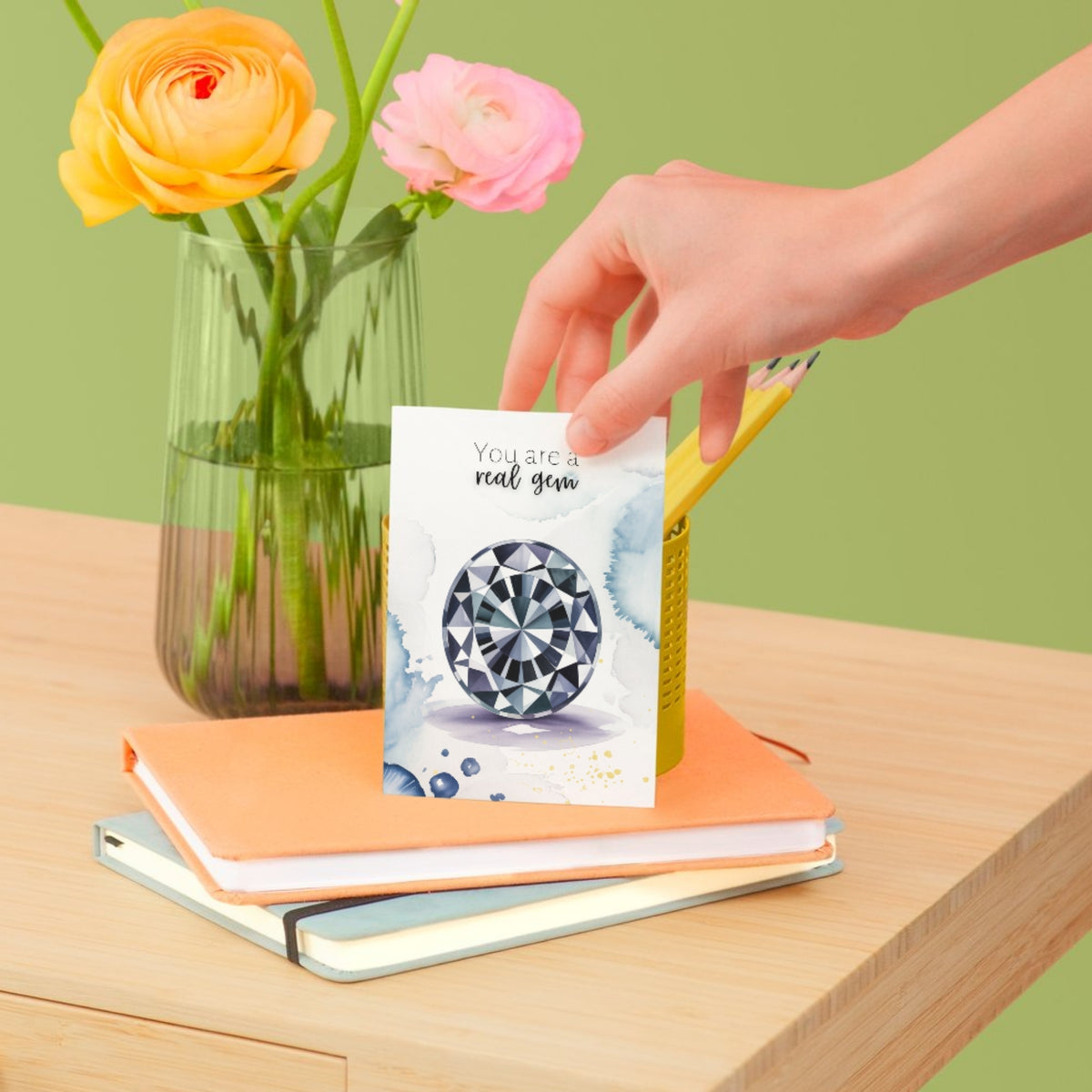 Hand holding a folded Gemstone greeting card with flowers and books in the background