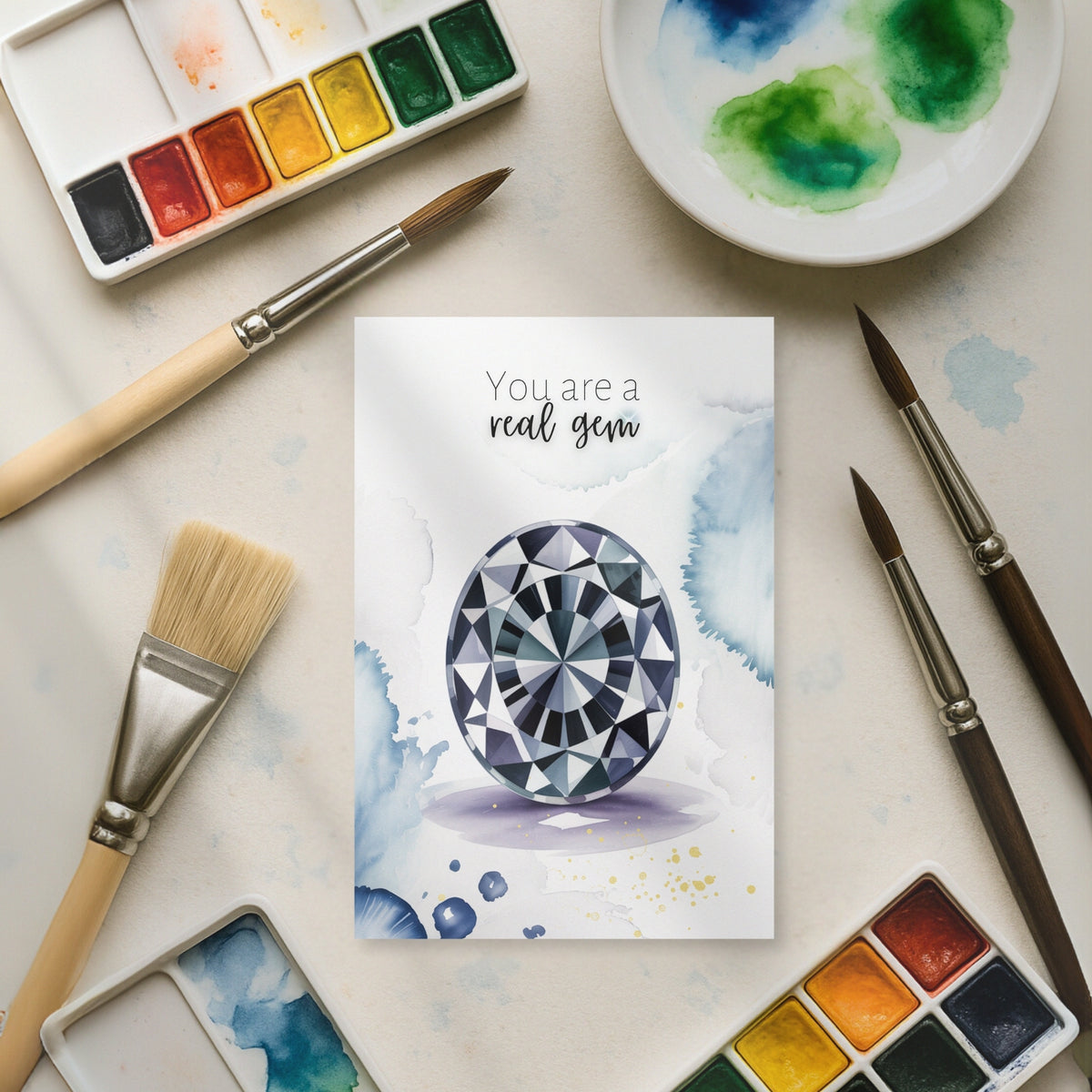 Card with watercolor design of a oval Spinel and text 'You are a real gem' surrounded by watercolor paints and brushes.