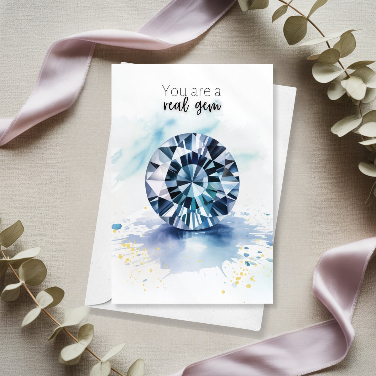 Greeting card with a blue spinel gemstone illustration and text 'You are a real gem' on a beige surface with ribbons and leaves.