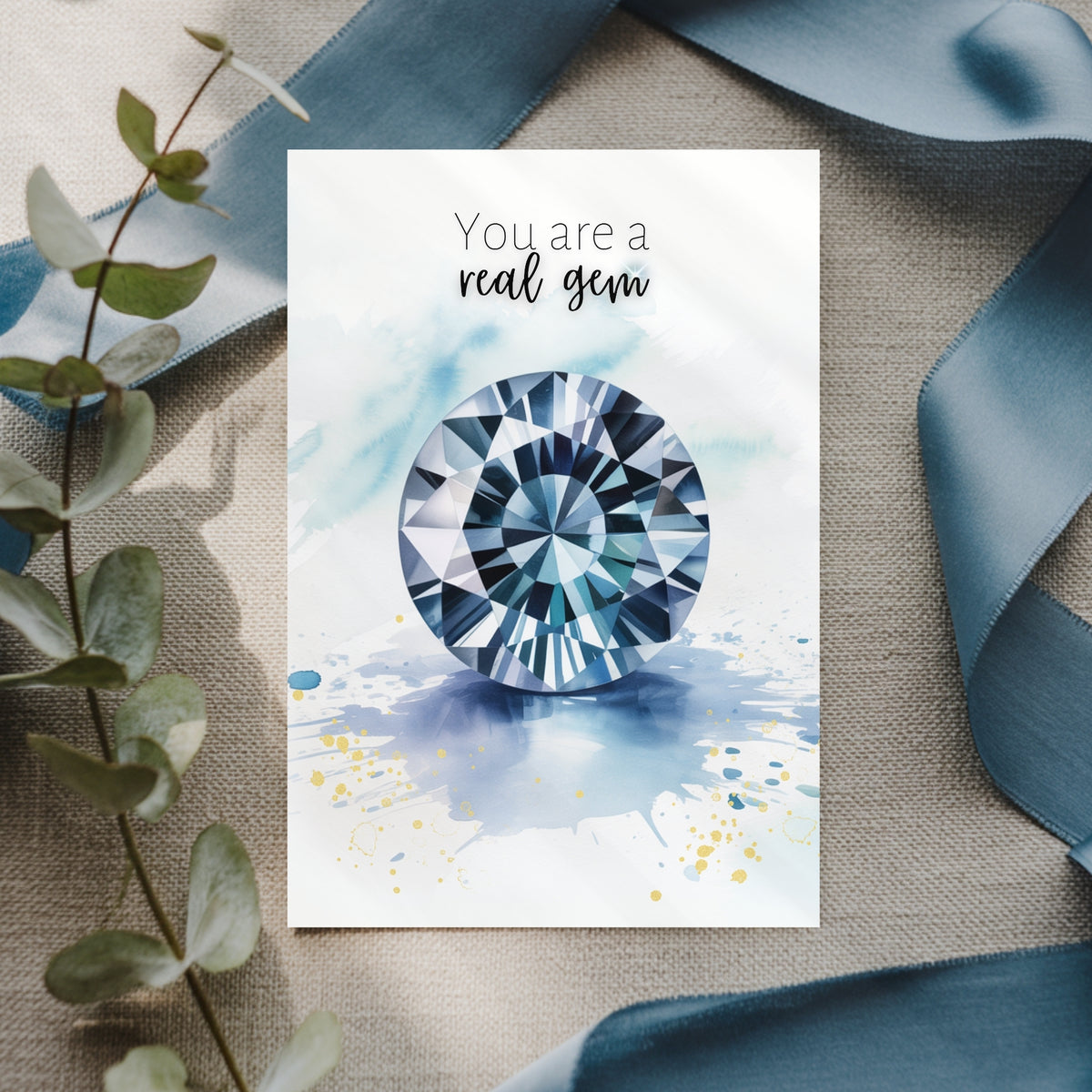Greeting card with a blue spinel gemstone illustration and text 'You are a real gem' on a textured surface with ribbons and leaves.