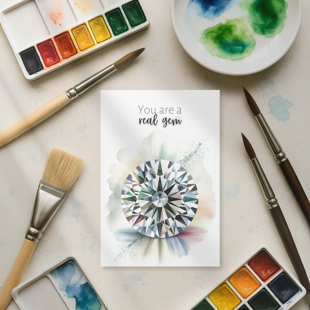Artistic card with diamond illustration surrounded by watercolor paints and brushes on a light surface.