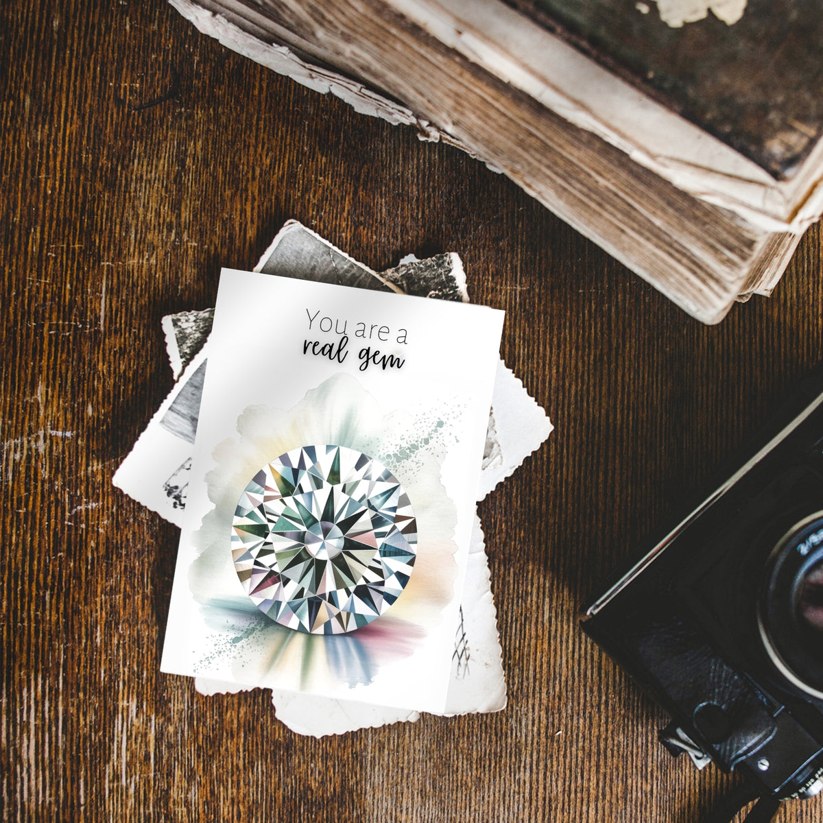 Card with diamond watercolor illustration and 'You are a real gem' text on a wooden surface with books and a camera.