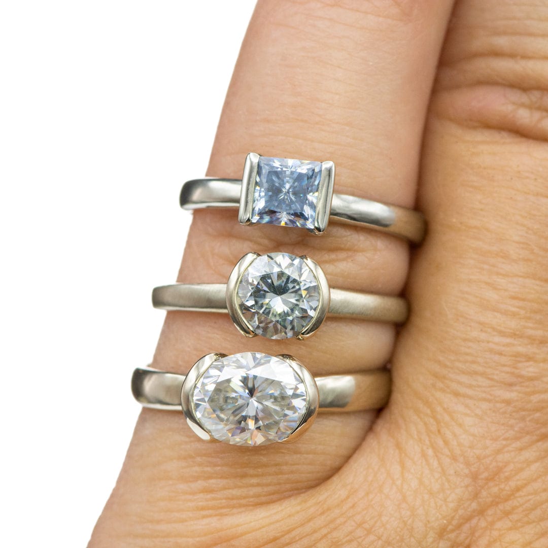 Three white gold rings with Moissanite gemstones on a person's finger, displayed against a white background. The top ring has a square-cut stone, the middle a round-cut stone, and the bottom a larger oval-cut stone, all in half bezel settings.Rings by Nodeform