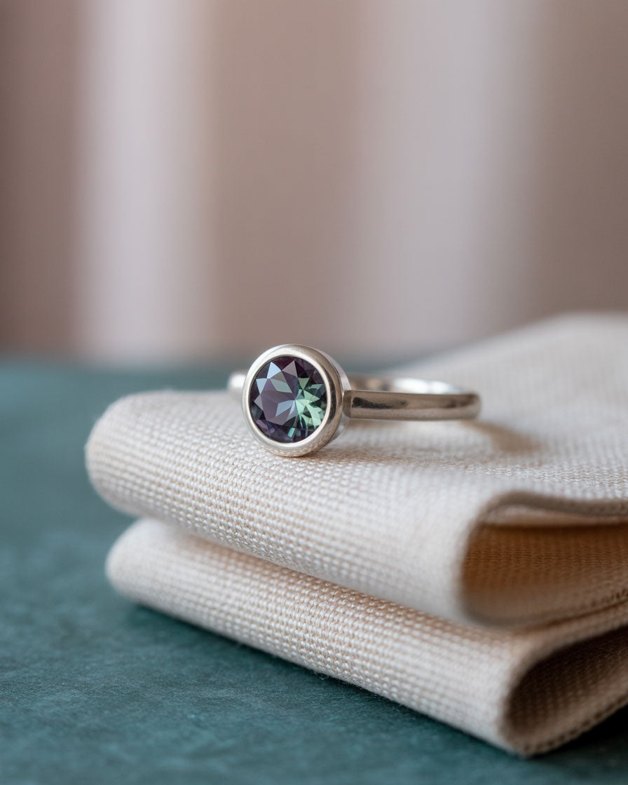 modern round alexandrite bezel set solitaire engagement ring on beige fabric
