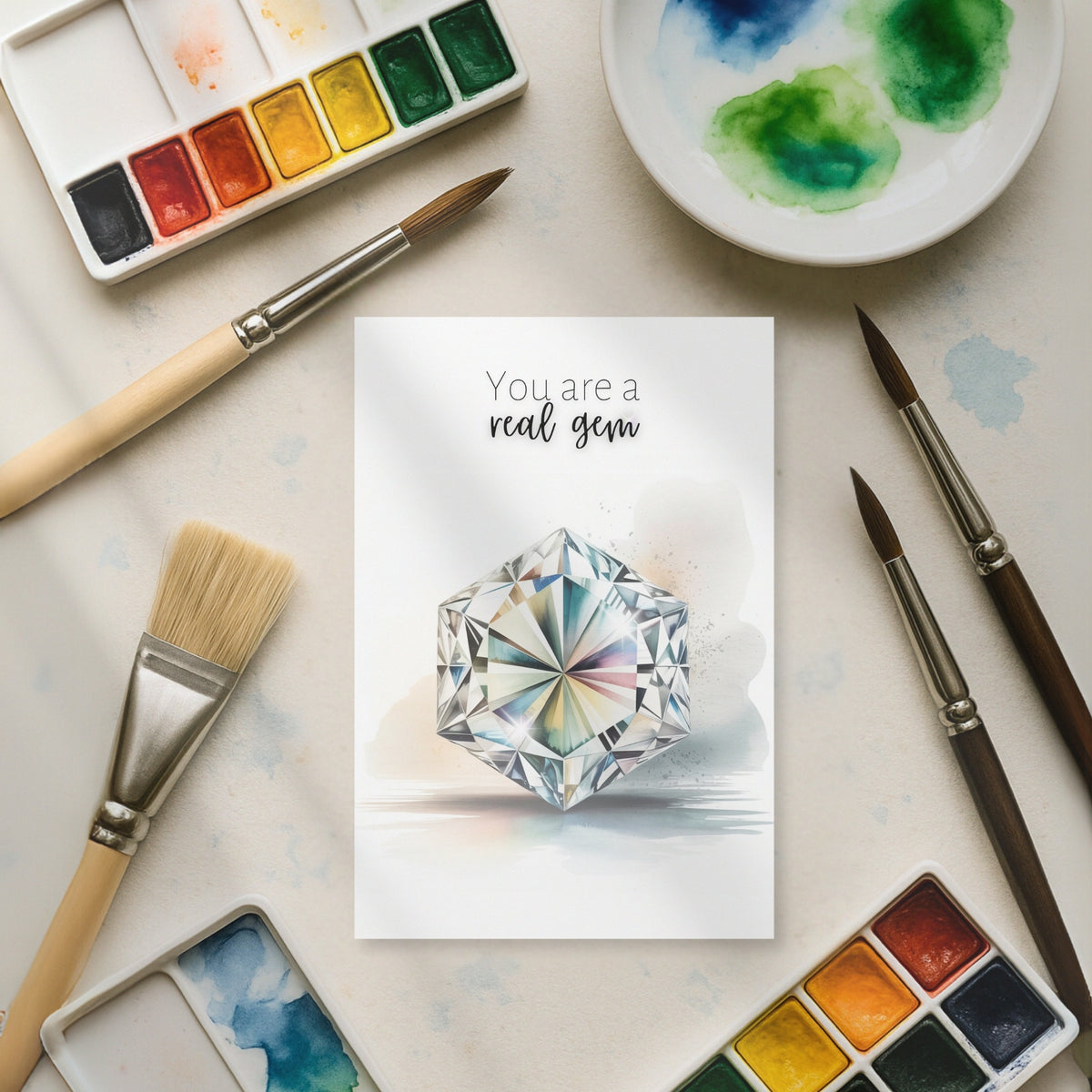 Card with a hexagon diamond illustration surrounded by watercolor paints and brushes on a light surface.