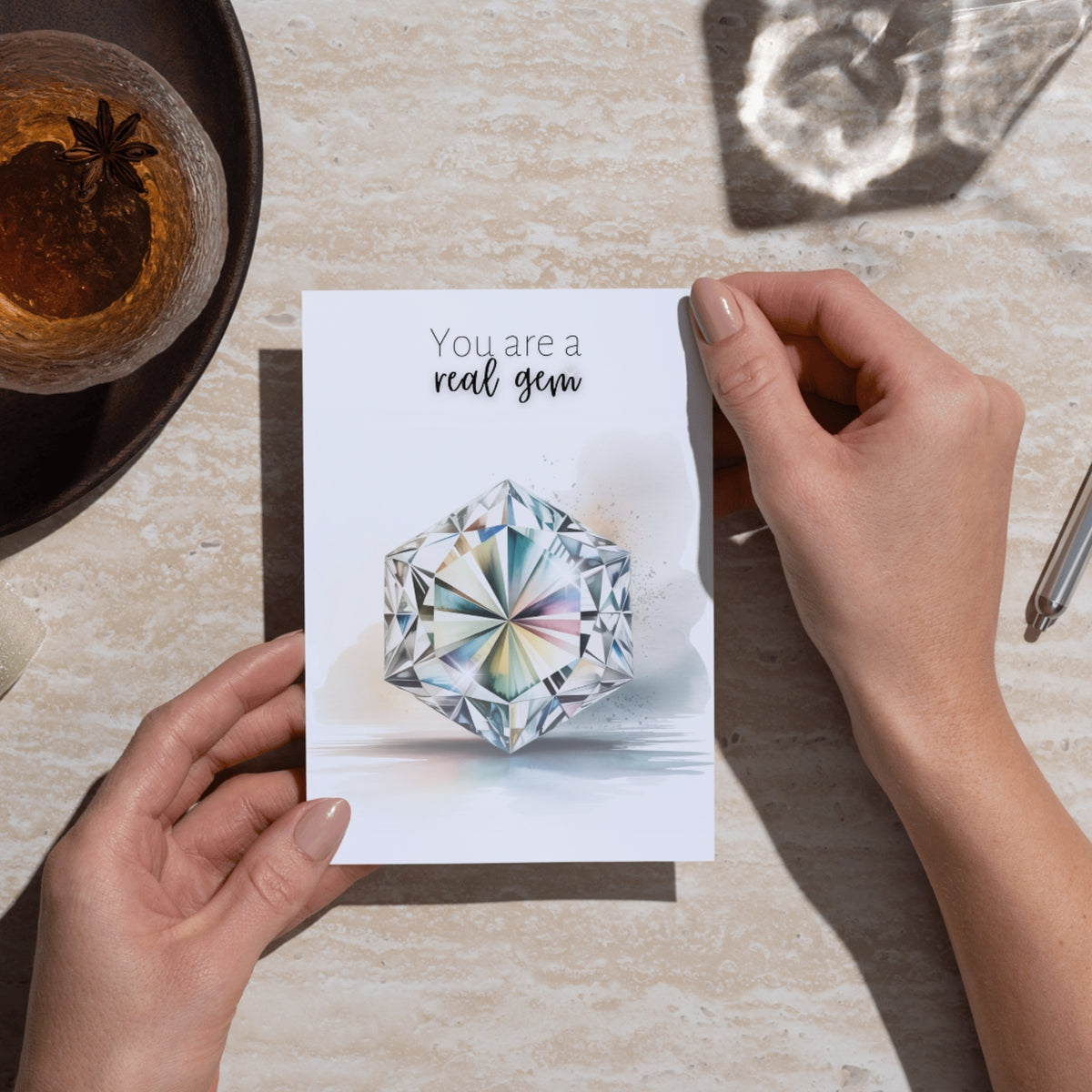Person holding a card with a hexagon diamond illustration and 'You are a real gem' text on a table.