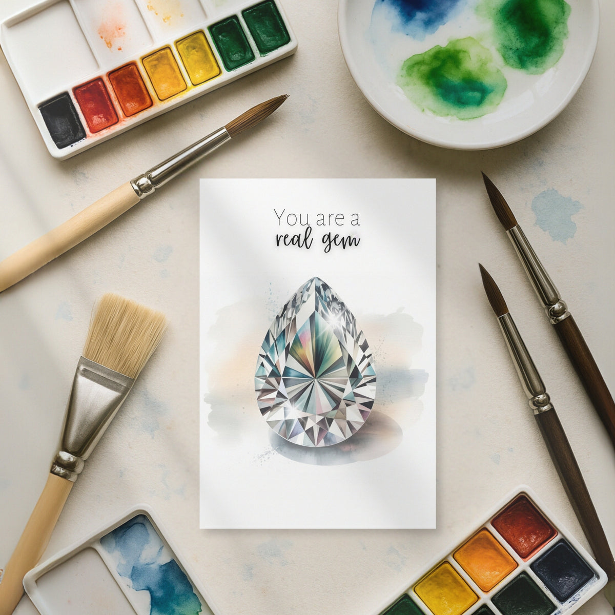 Card with a diamond illustration surrounded by watercolor paints and brushes on a light surface.