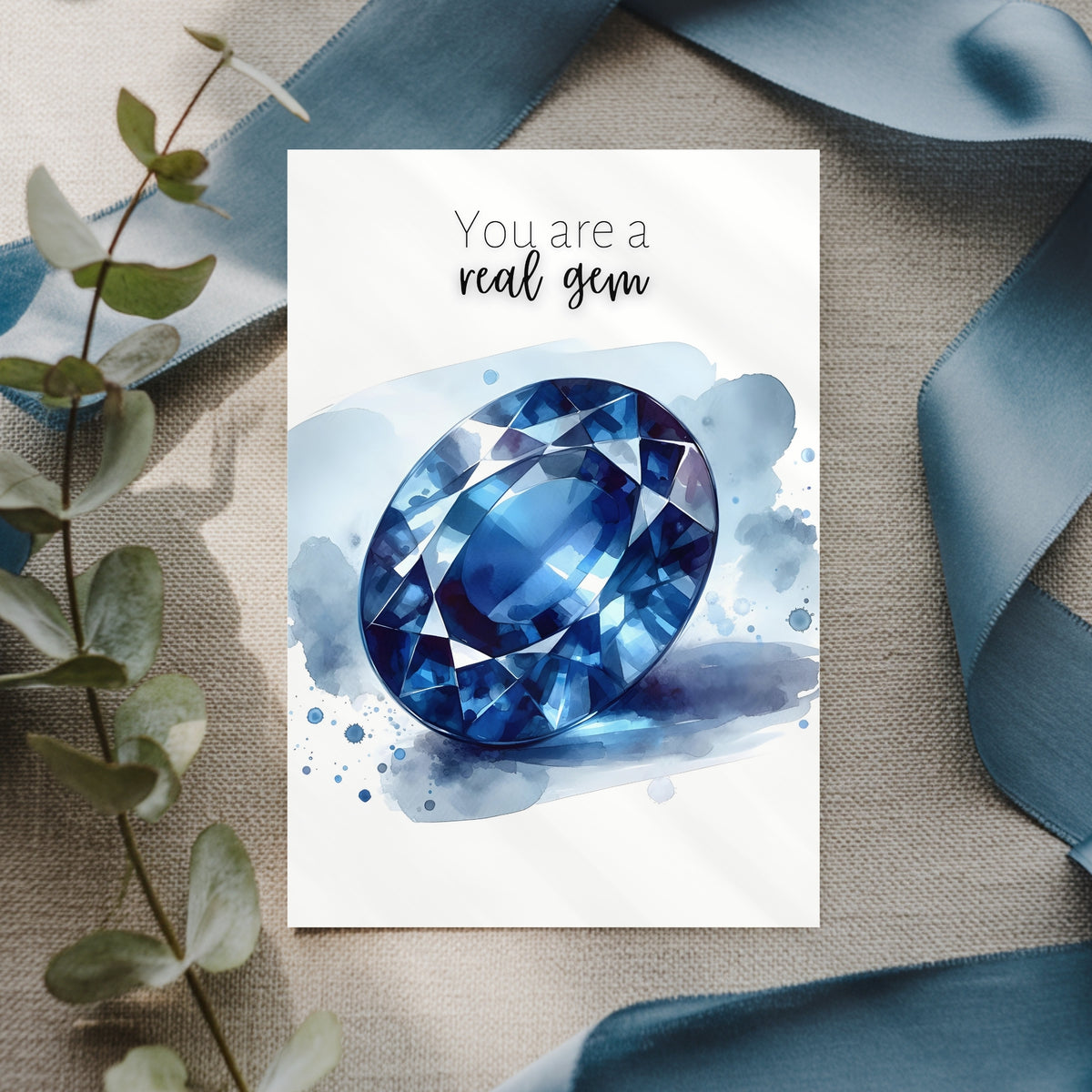 Card with blue sapphire watercolor illustration and text 'You are a real gem' on a textured surface with decorative elements.