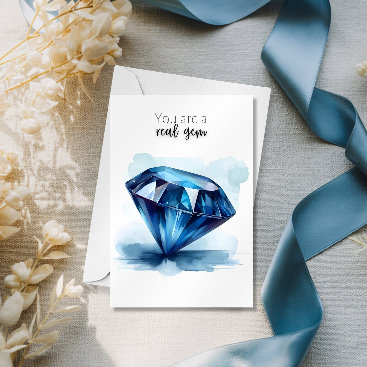 Greeting card with blue sapphire watercolor illustration and text 'You are a real gem' on a textured surface with flowers and ribbon.