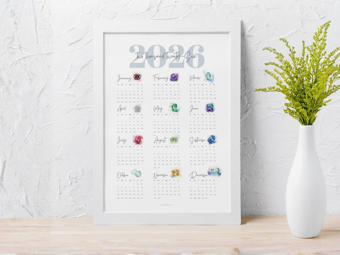 Framed 2026 birthstone calendar with colorful gemstones on a white textured wall background.