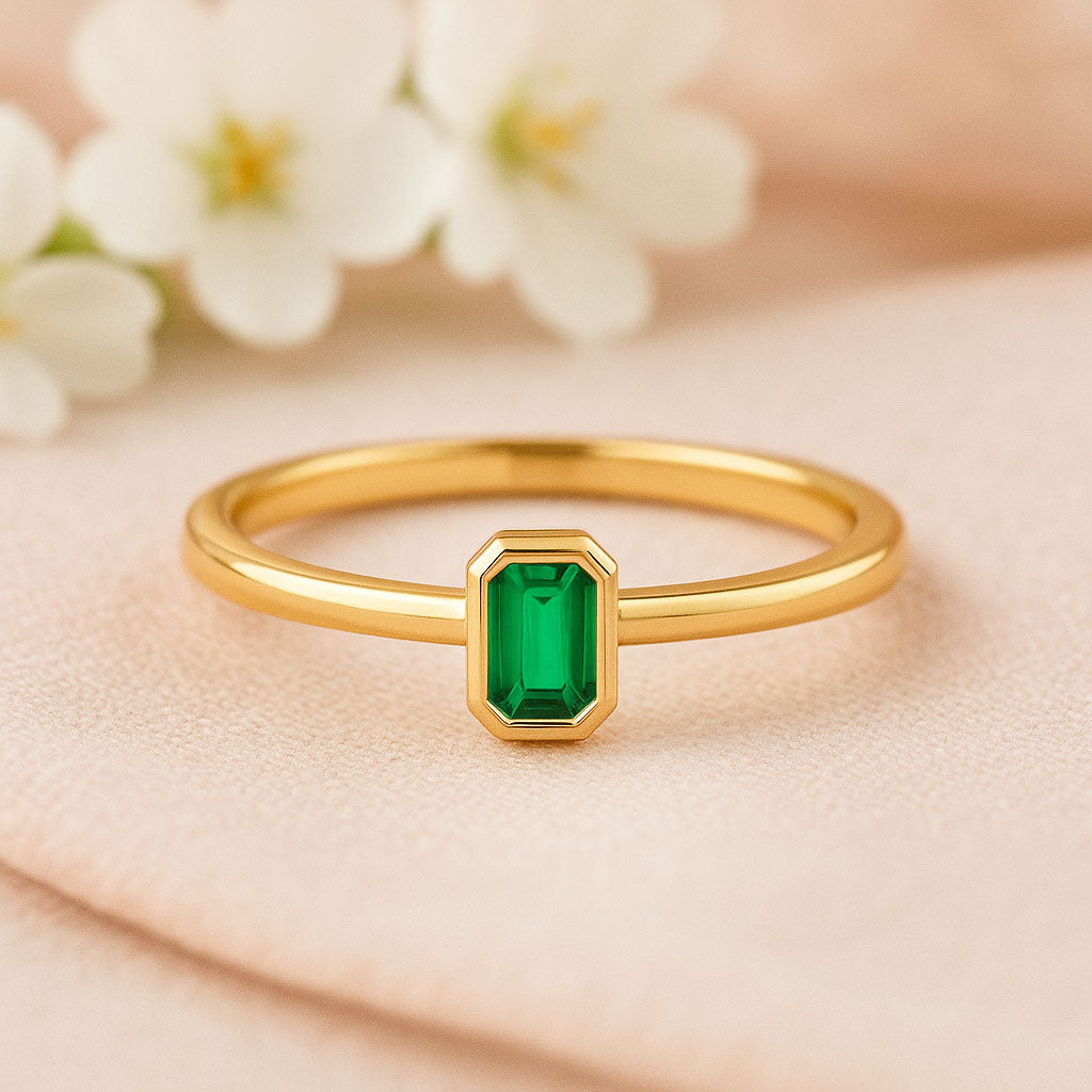 Minimalist gold ring with a bezel set emerald cut green lab emerald stone, resting on a light-colored cloth.