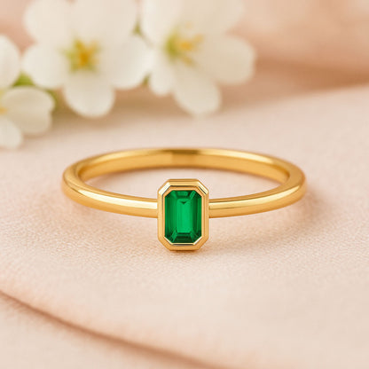 Minimalist gold ring with a bezel set emerald cut green lab emerald stone, resting on a light-colored cloth.