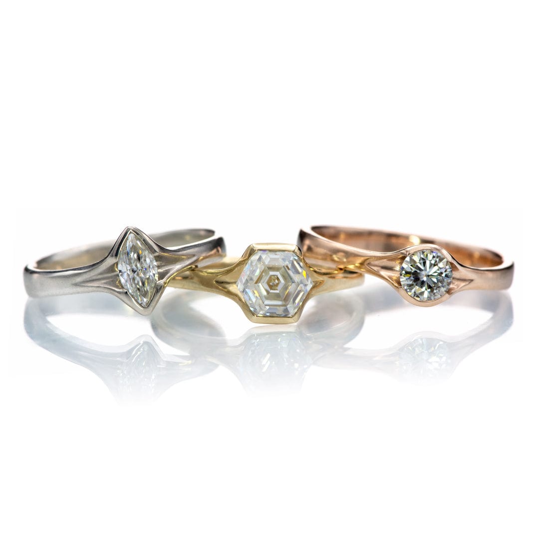 Three rings with a marquise, hexagon amd round moissaniteare displayed against a white background with their reflections visible on the surface. 